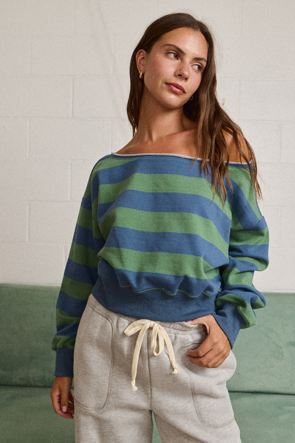 A woman wearing a blue and green striped off-the-shoulder knit sweater and light gray sweatpants, standing in front of a white brick wall and a green couch.