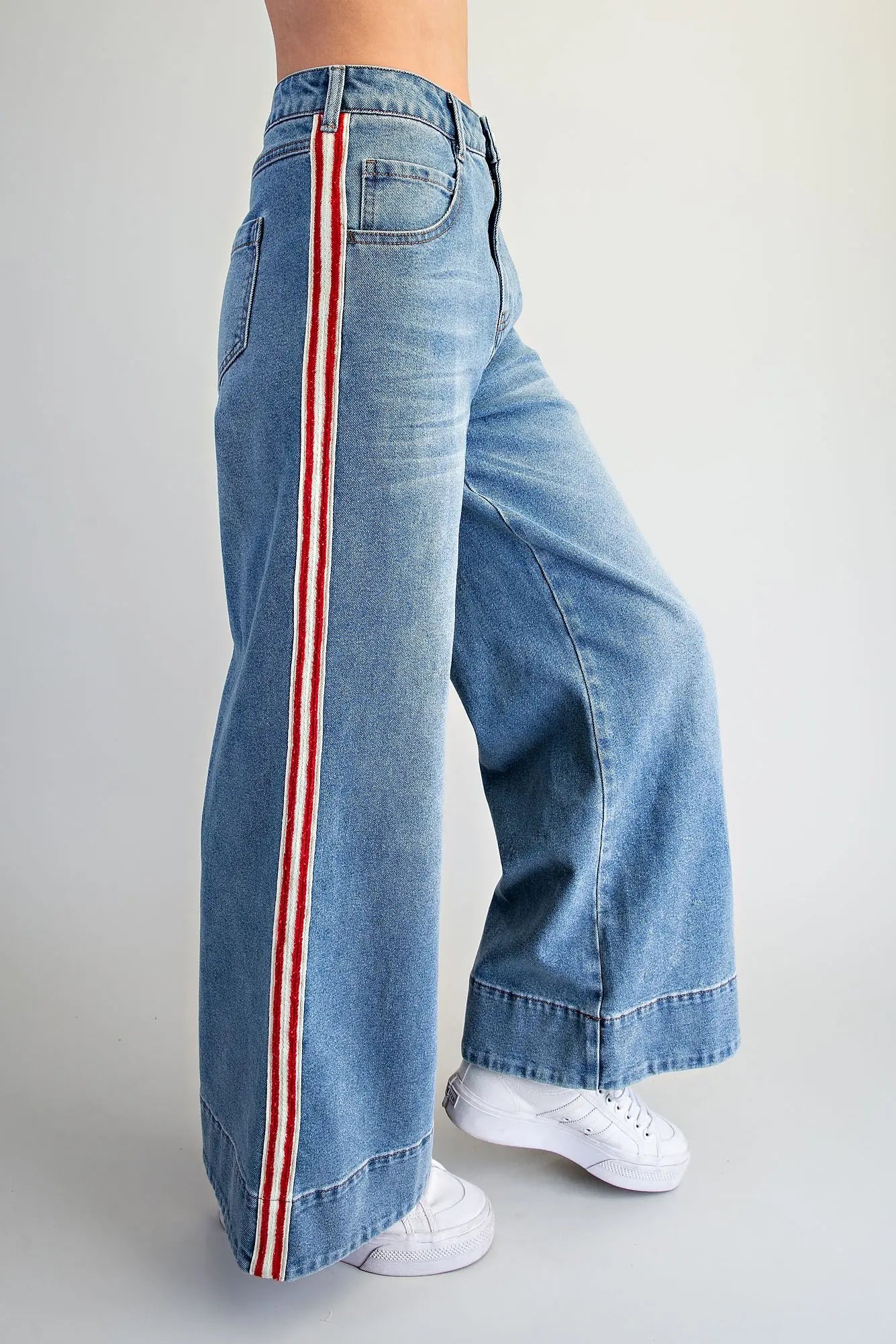 A person wearing light blue wide-leg denim jeans with a red and white vertical stripe down the side, paired with white sneakers, shown against a plain light gray studio background.