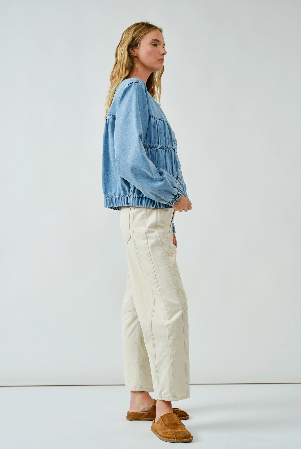 A women wearing a denim bomber jacket standing sideways beige pants and tan slides