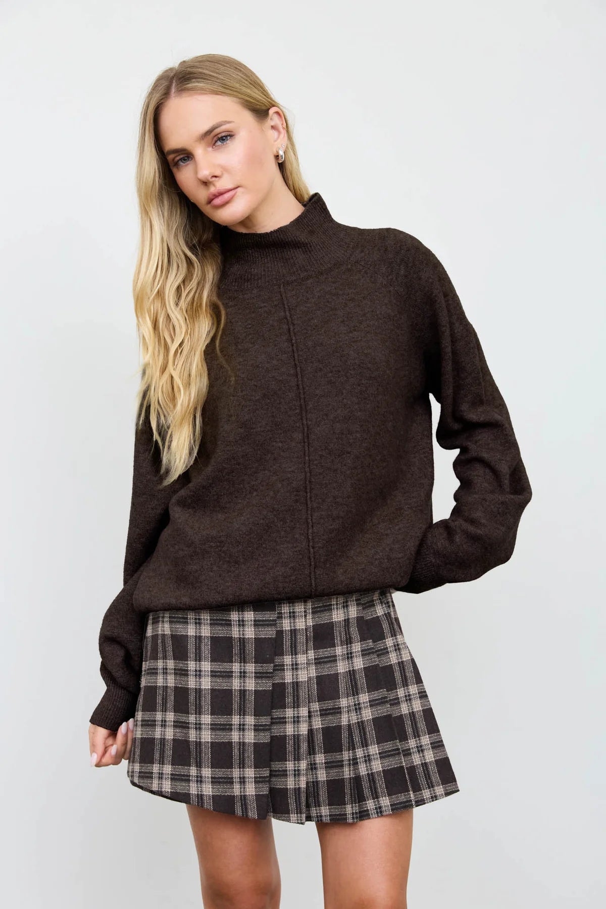 A woman modeling a dark brown mock-neck knit sweater paired with a brown and beige plaid pleated mini skirt, set against a plain light gray studio background.