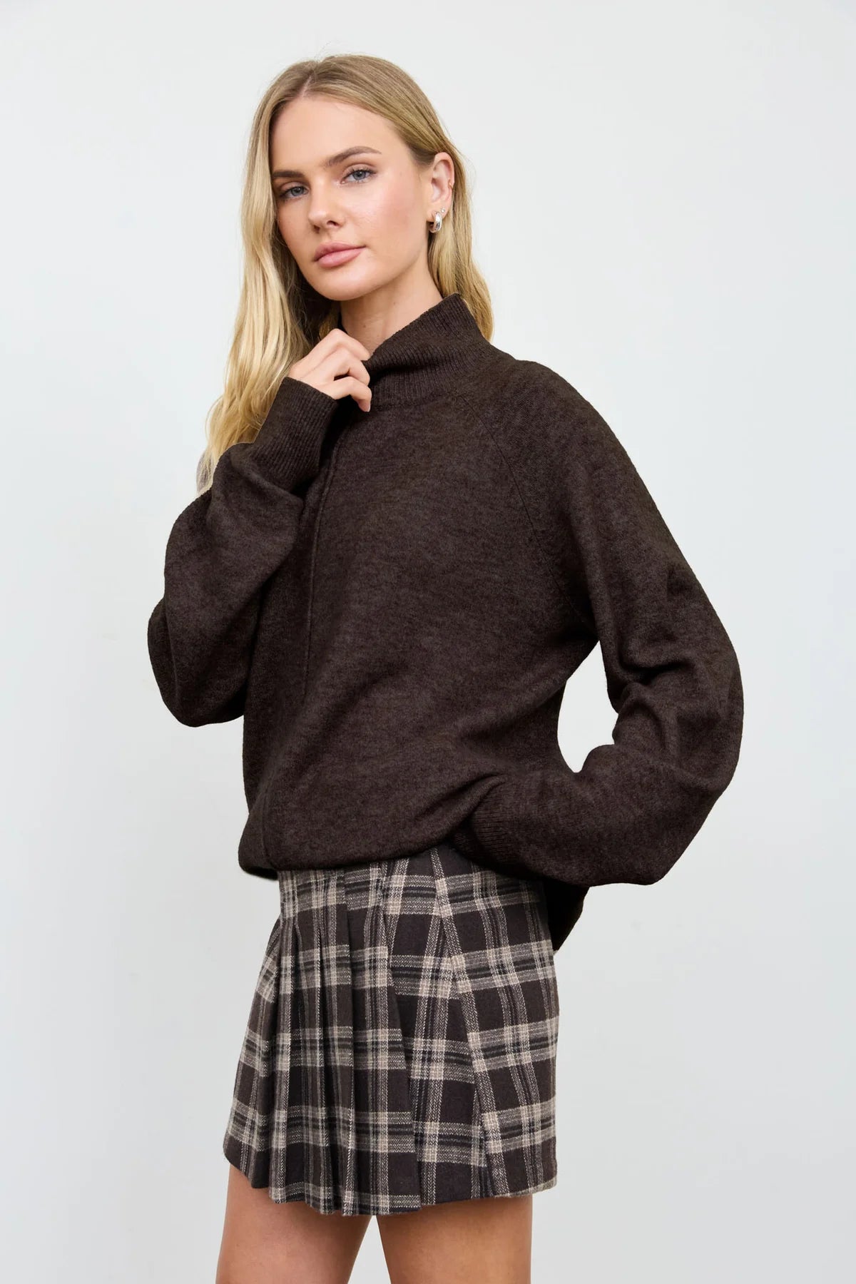 A woman modeling a dark brown mock-neck knit sweater paired with a brown and beige plaid pleated mini skirt, set against a plain light gray studio background.