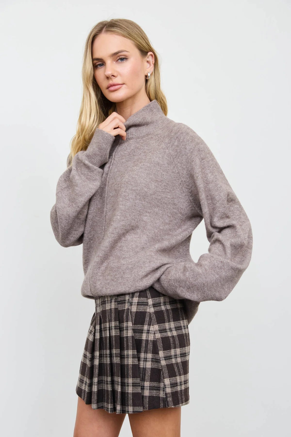 A blonde model wearing a heathered mocha-colored mock-neck knit sweater and a dark brown and beige plaid pleated mini skirt, posing against a light gray studio background.