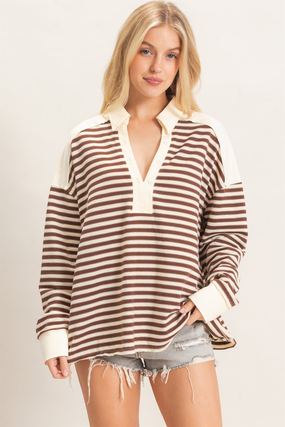A woman modeling a brown and cream horizontally striped long-sleeve henley sweatshirt with solid cream shoulder panels and collar, paired with distressed denim shorts against a light beige studio background.