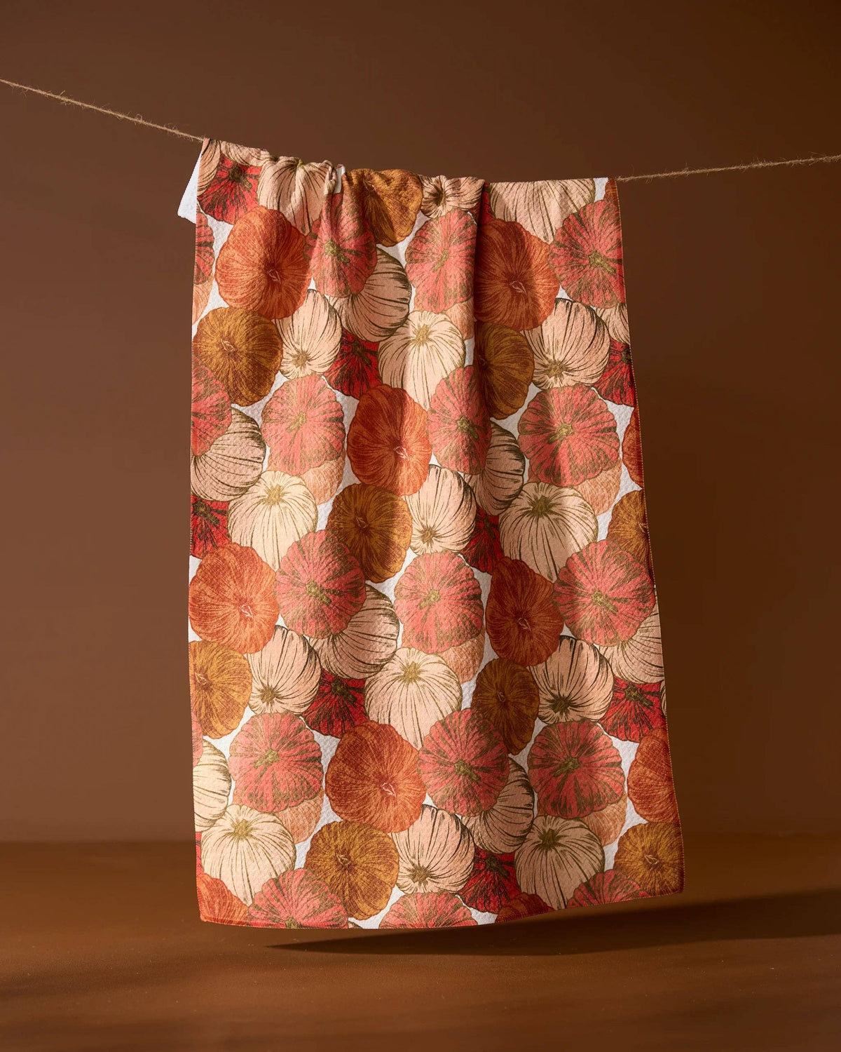 A white kitchen towel with a large, repeating autumnal pumpkin pattern in shades of red, orange, and beige, hanging from a thin rope against a warm brown background.