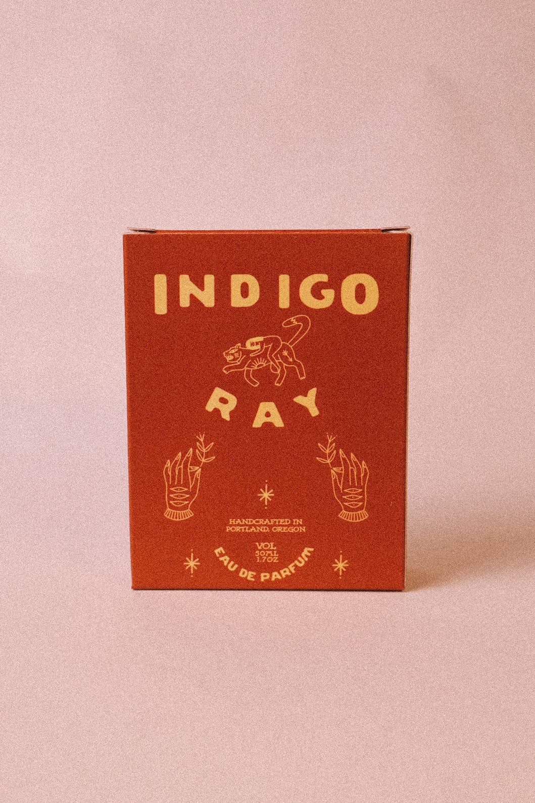 A square glass bottle of Indigo Eau de Parfum with a black cap and a cream-colored label featuring a dark red logo, set against a solid light pink background.