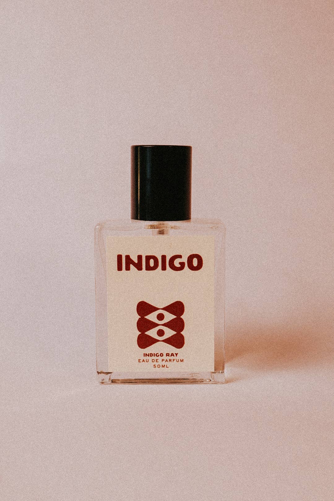 A square glass bottle of Indigo Eau de Parfum with a black cap and a cream-colored label featuring a dark red logo, set against a solid light pink background.