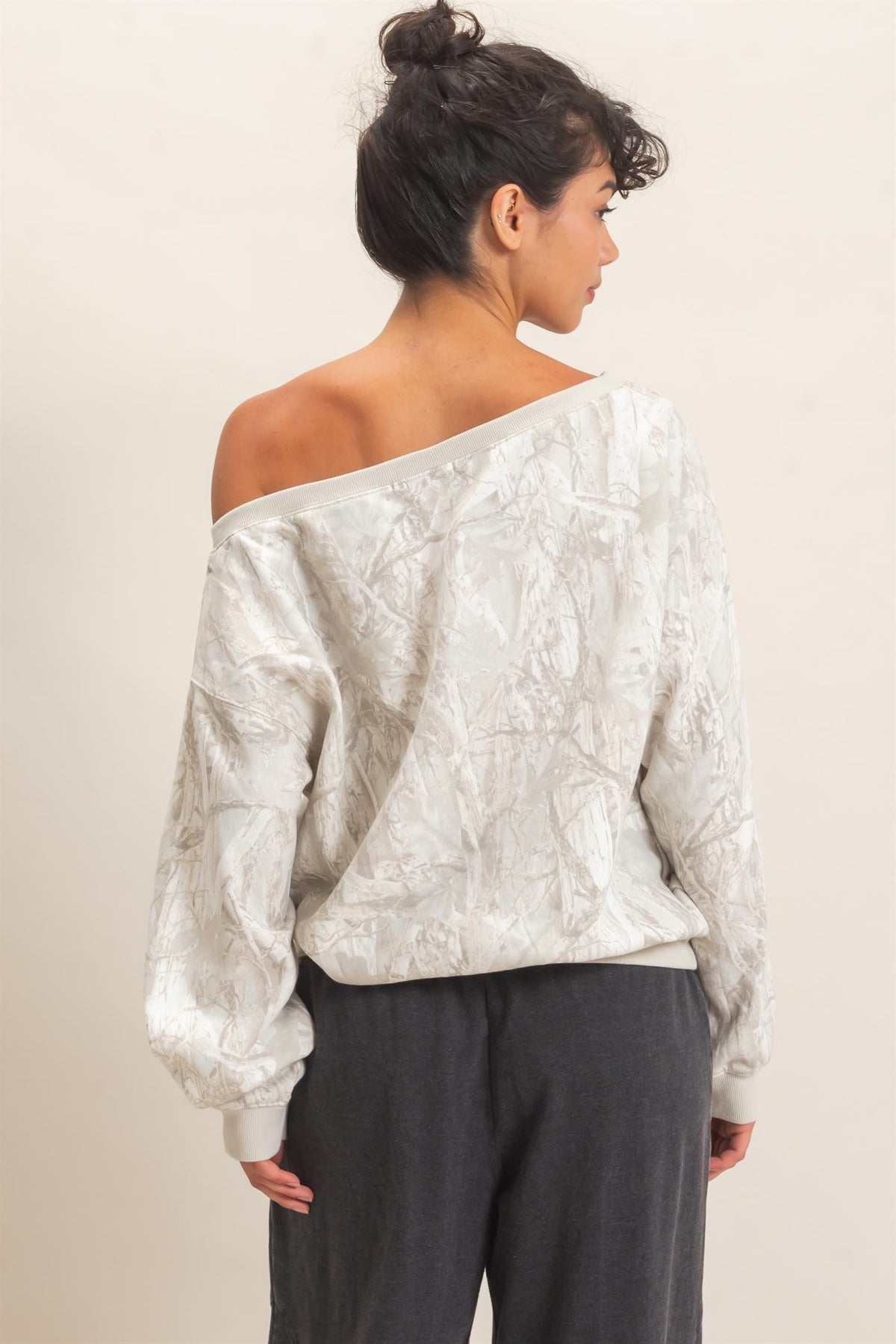 A woman shown from the back wearing a textured off-the-shoulder long-sleeve top in a light cream or off-white color, paired with dark gray pants, against a plain light beige background.