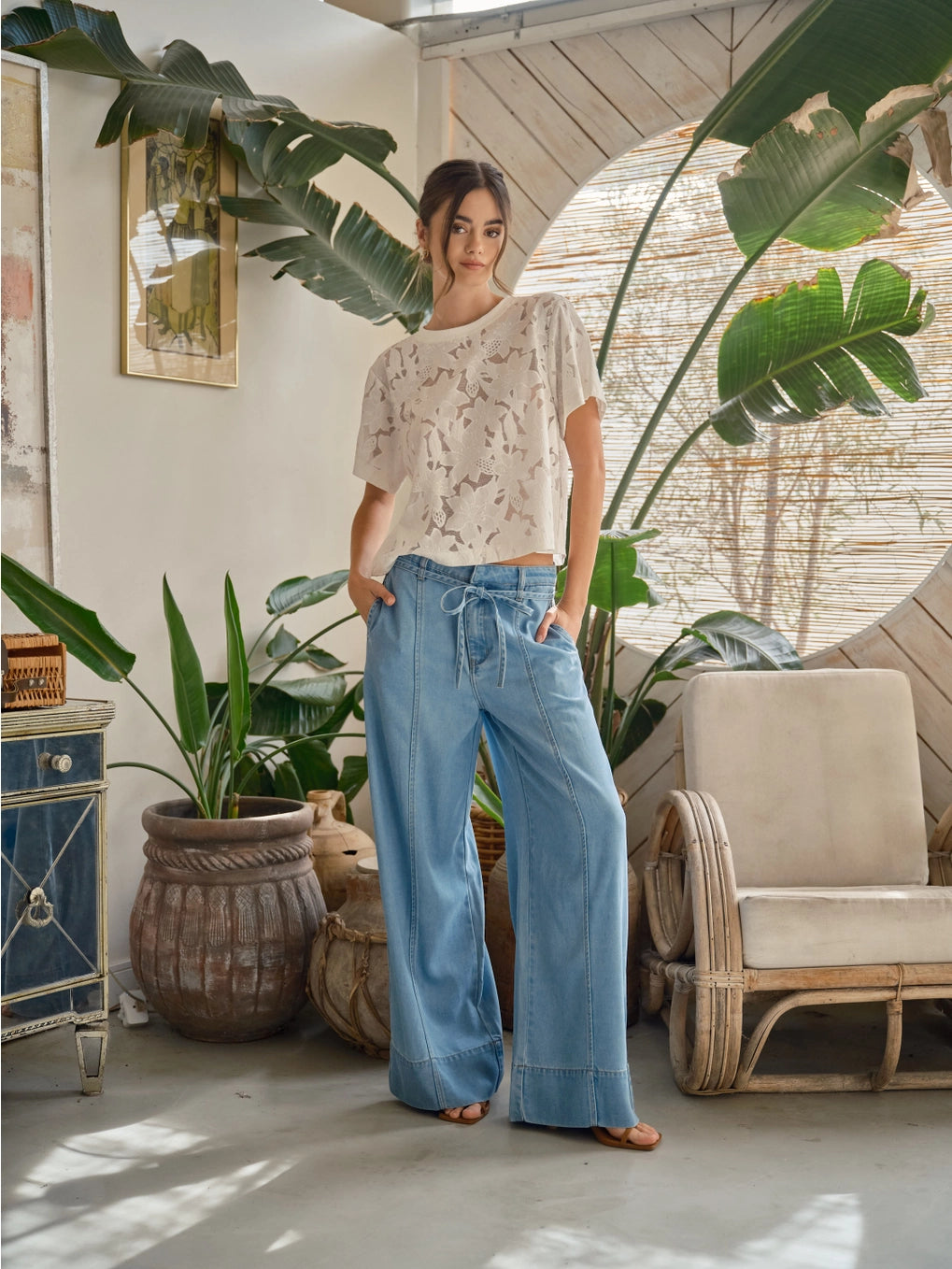 Woman standing in a room with plants and furniture wearing a off white lace floral t-shirt.