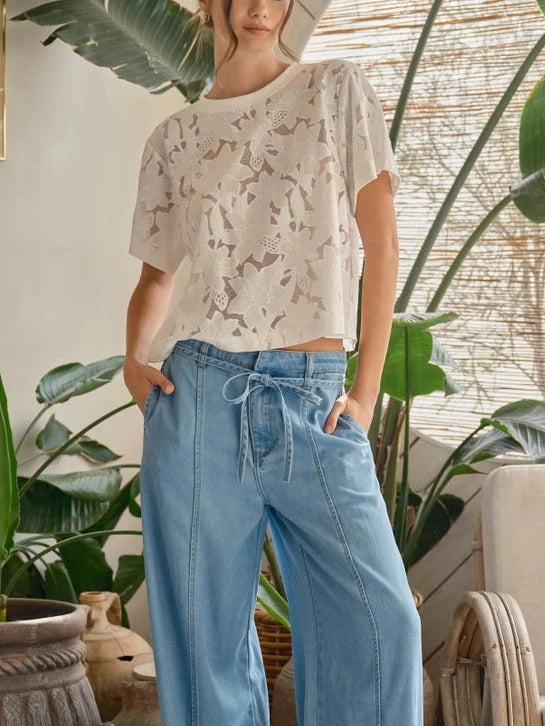 Woman wearing a white lace floral top and blue jeans against a plain background